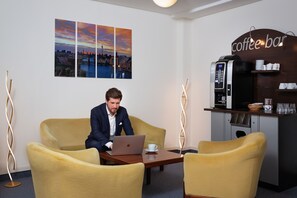 En mand sidder ved et bord med en bærbar computer, to gule stole, en kaffemaskine og et bybillede på væggen.