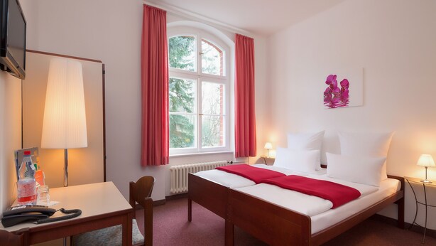 A hotel room with a bed, bedside tables, a desk, a chair, a window with curtains, and a wall-mounted artwork. - Hotel Morgenland (Berlin)