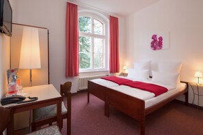 Et hotelværelse med seng, sengeborde, et skrivebord, en stol, et vindue med gardiner og et billede hængende på væggen.