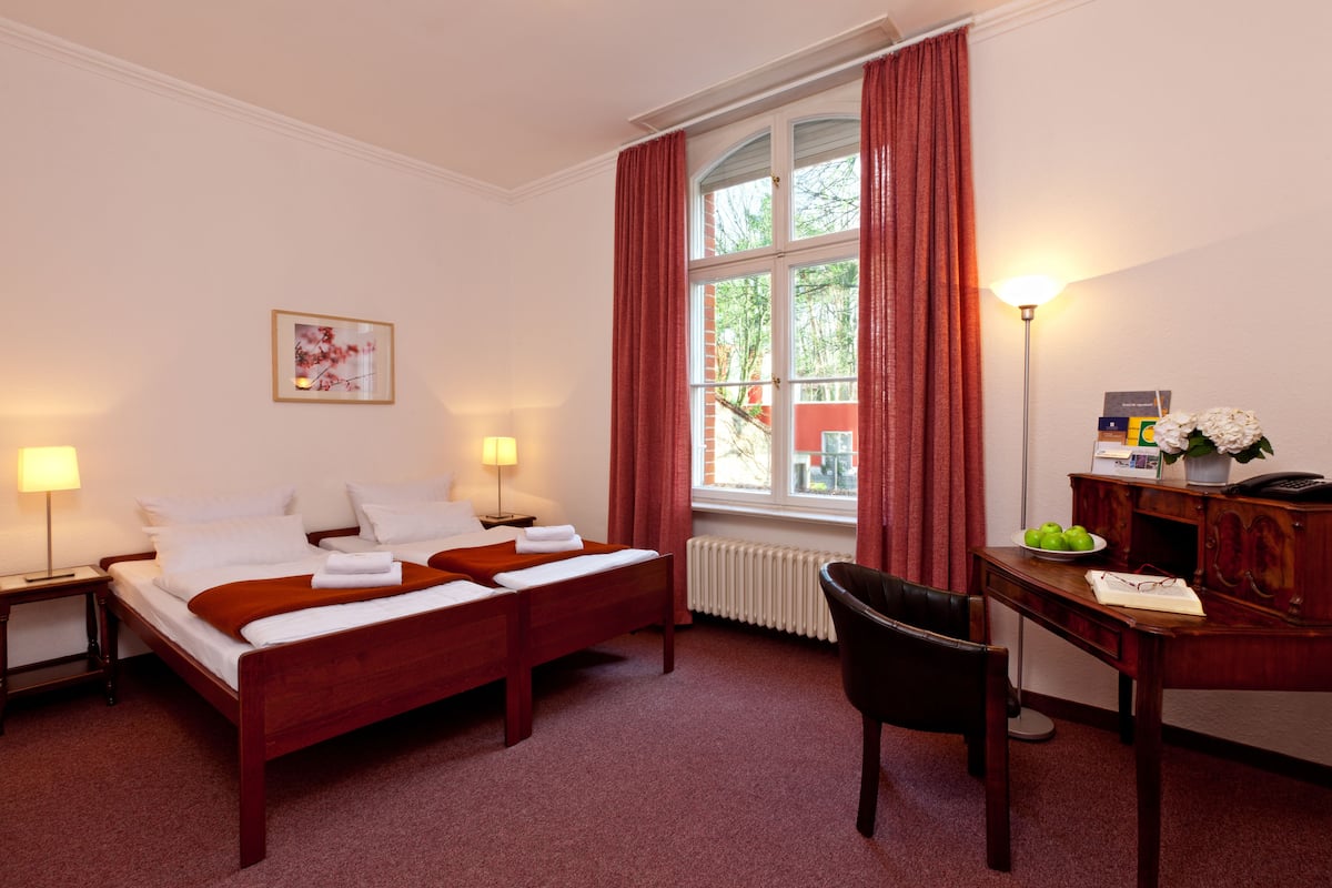 Quarto de hotel com cama de casal, duas mesas de cabeceira com luminárias, escrivaninha com cadeira, radiador e janela com cortinas.
