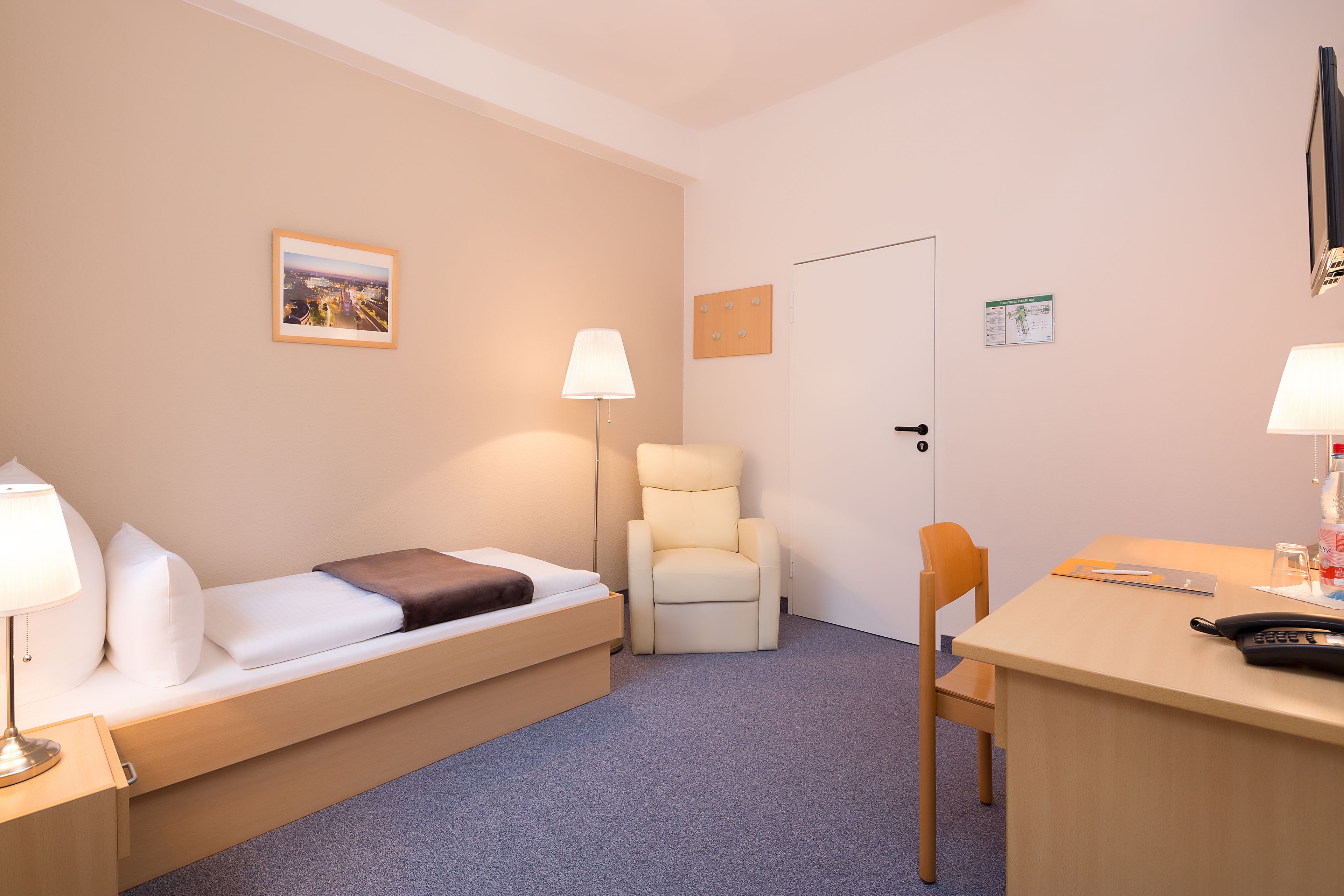 A compact hotel room with a single bed, a chair, a desk, and a lamp.