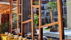 Un espacio moderno al aire libre con sofás amarillos y mesas de madera, amplios ventanales y vistas a un edificio con balcones.