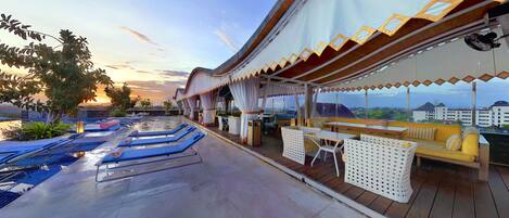 A rooftop lounge area with sun loungers, a seating area with cushions, and a view of the cityscape.