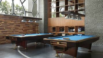 A pool table with a wooden finish and metal legs, a second pool table with a similar design, and a seating area with chairs and a table.