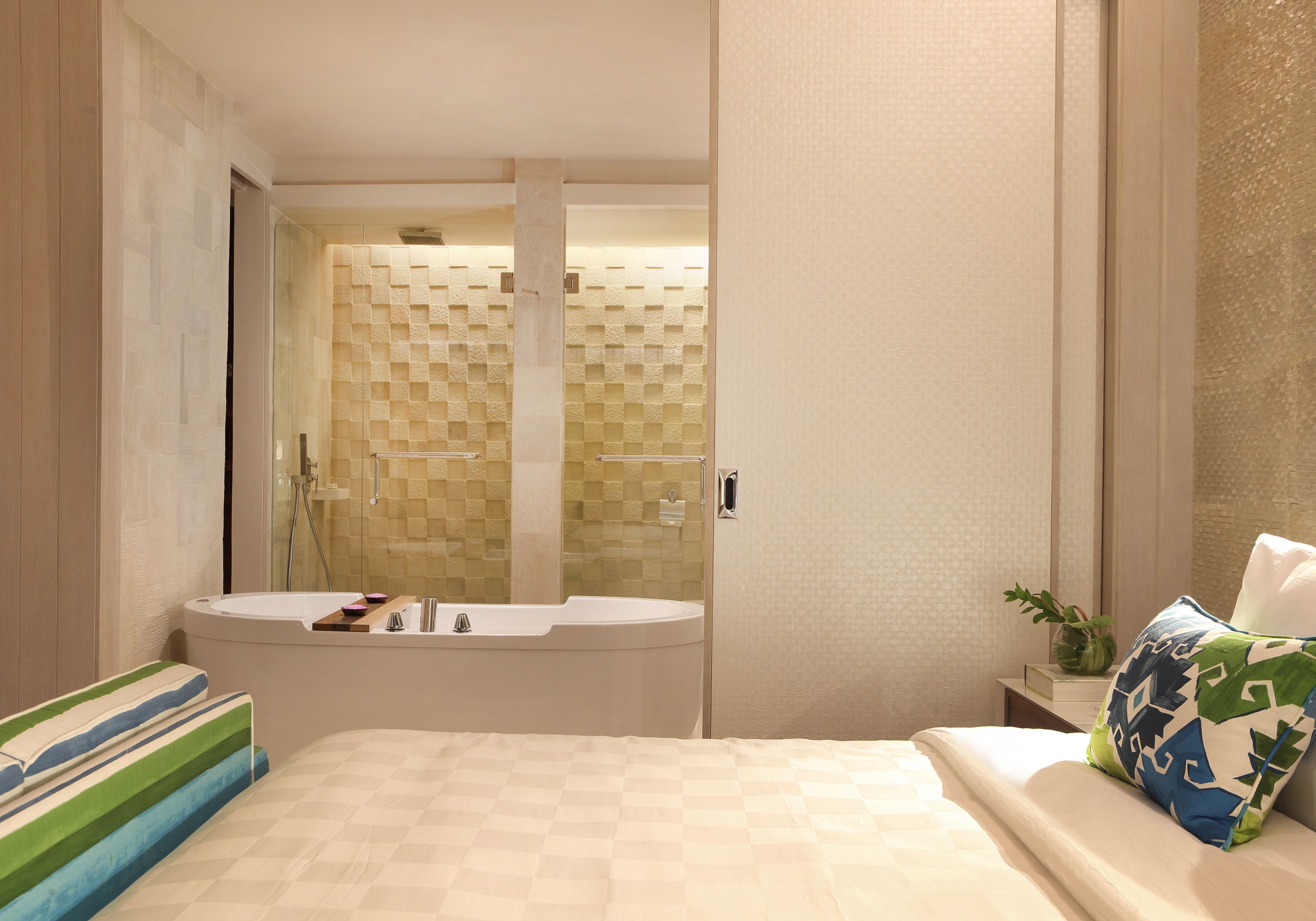 A hotel room with a large bathtub, a bed with striped pillows, and a small bedside table with a vase.