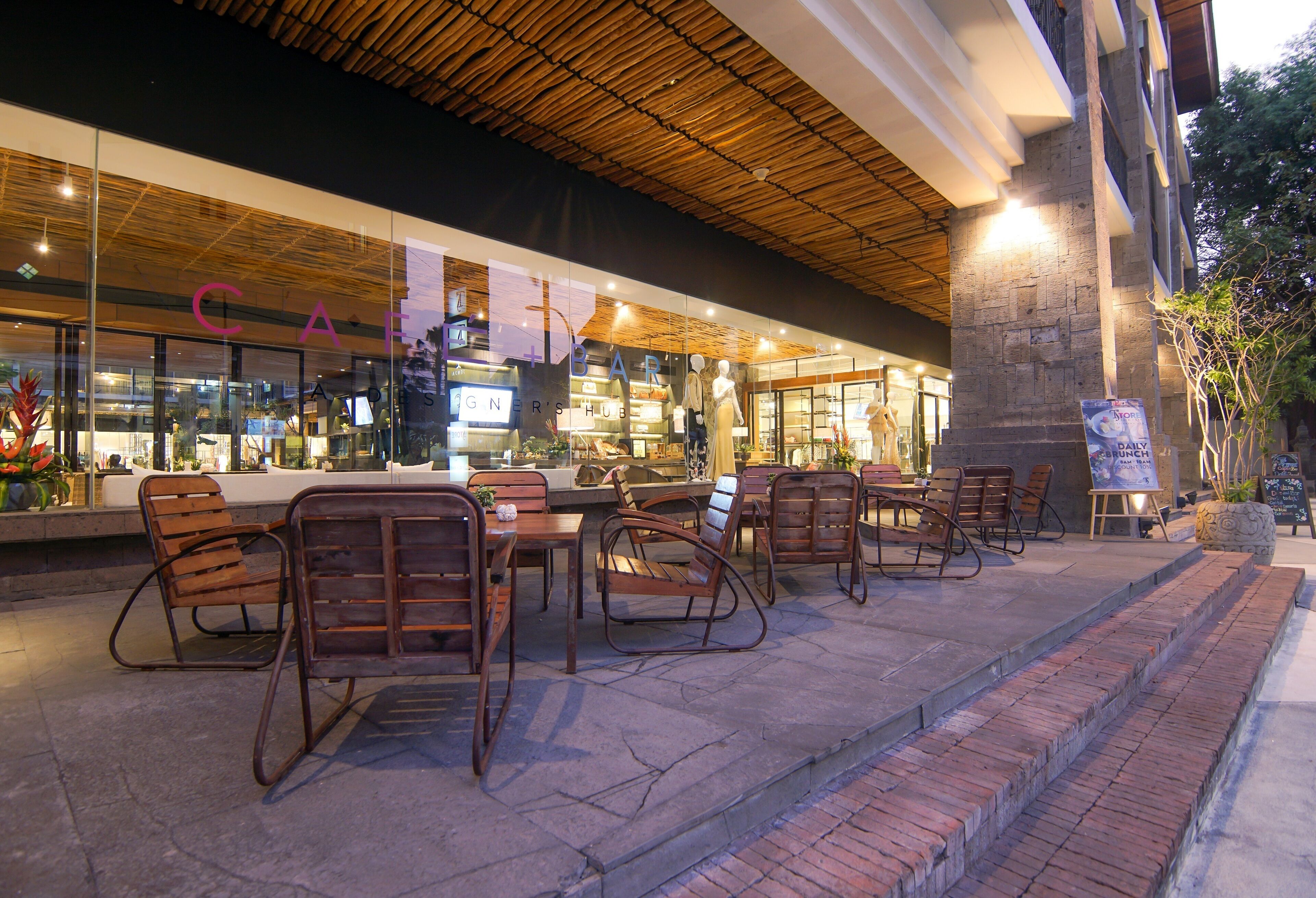 An outdoor seating area with wicker chairs and tables in front of a building with a glass facade and the word "CAFE" visible.