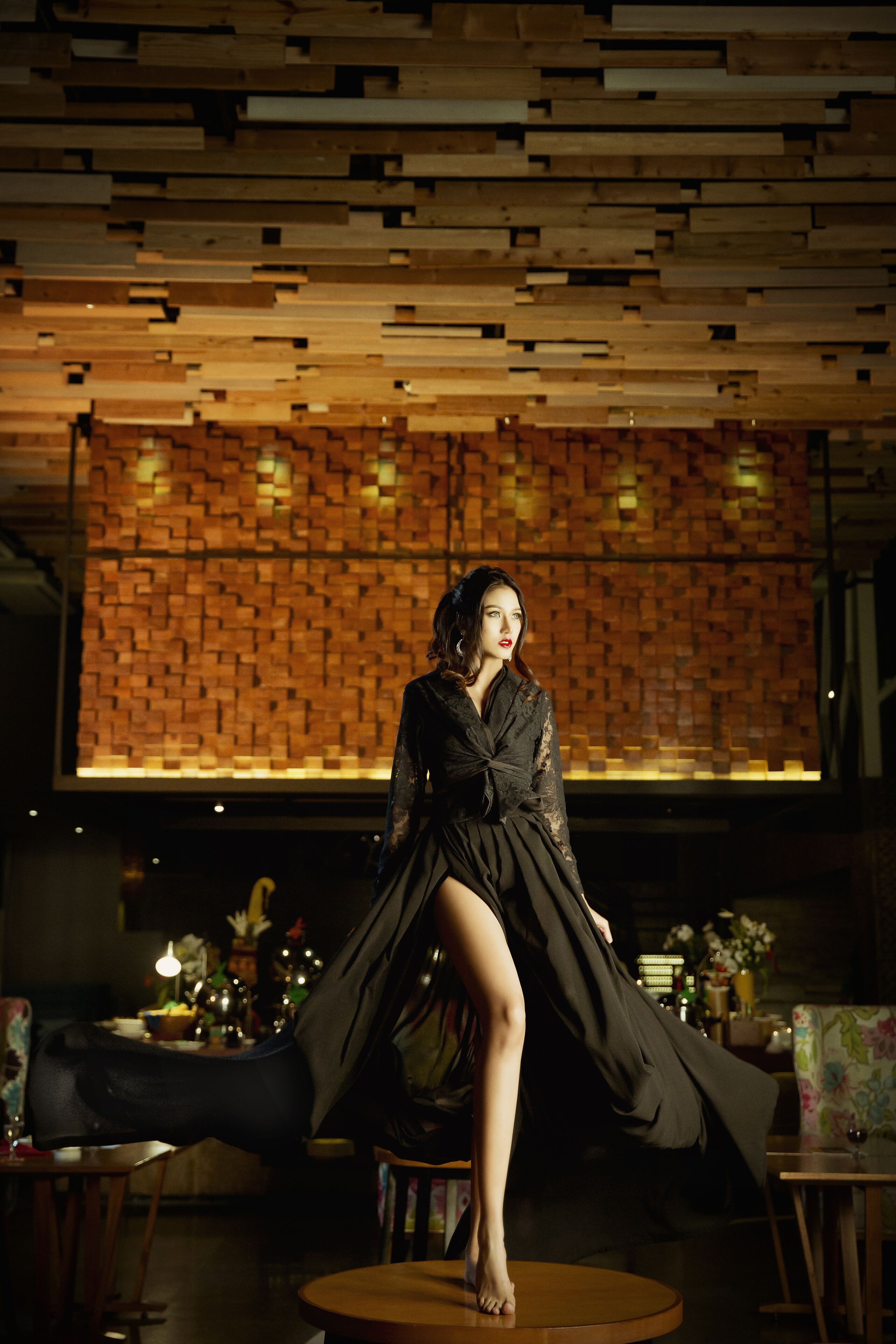 A woman seated on a round table in a dimly lit room with a brick-patterned wall and a modern ceiling design.