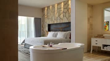A modern hotel room with a large bed, a built-in bathtub, a vanity with a mirror, and a stone accent wall.