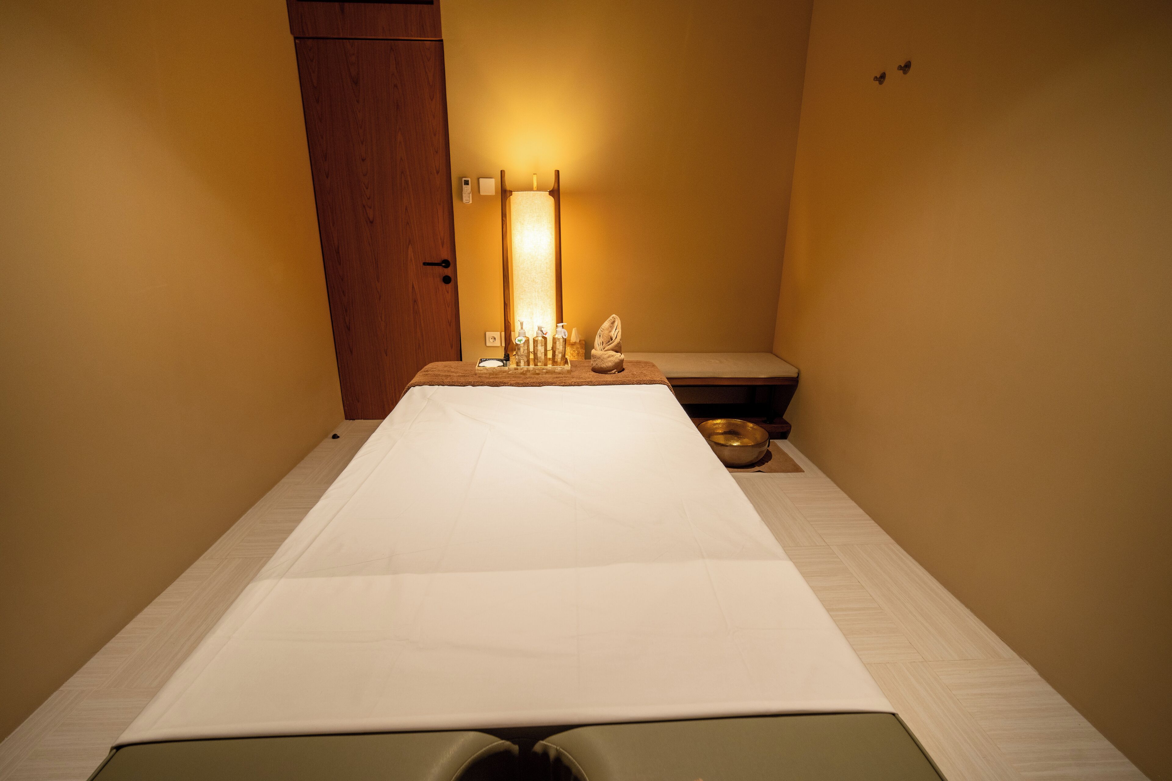 A room with a bed, a wooden door, a wall-mounted light fixture, and a small table with a bowl and a bottle.