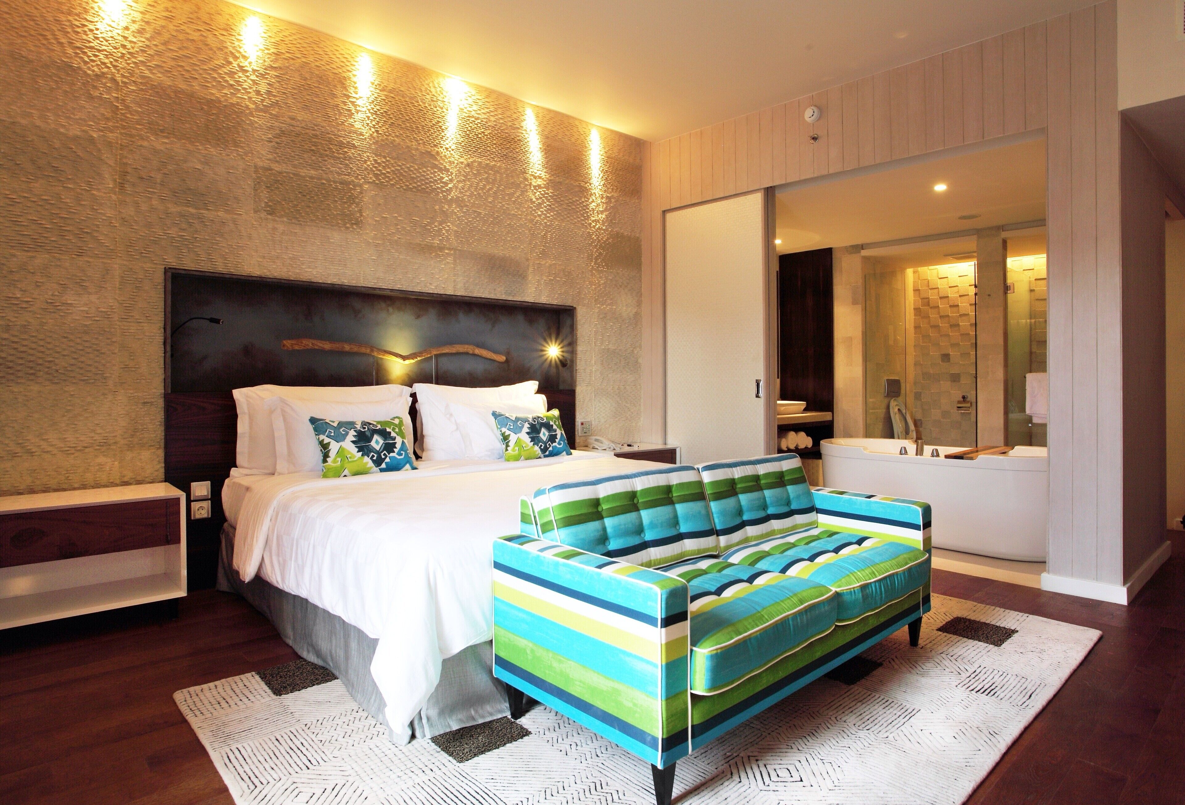A modern hotel room with a large bed, a colorful striped sofa, a large mirror, and a separate bathroom with a bathtub.