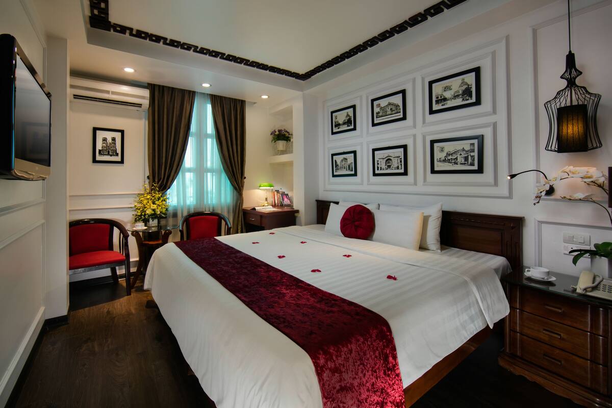 A hotel room with a large bed, a desk with a lamp, a chair, and framed pictures on the wall.