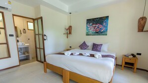 A bedroom with a large bed, a bedside table, a nightstand, a painting, and a bathroom visible through an open door. - Palm Garden Resort (Rawai)