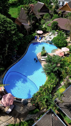 Une piscine où se trouvent deux personnes, entourée d’une végétation luxuriante et de quelques chaises longues.