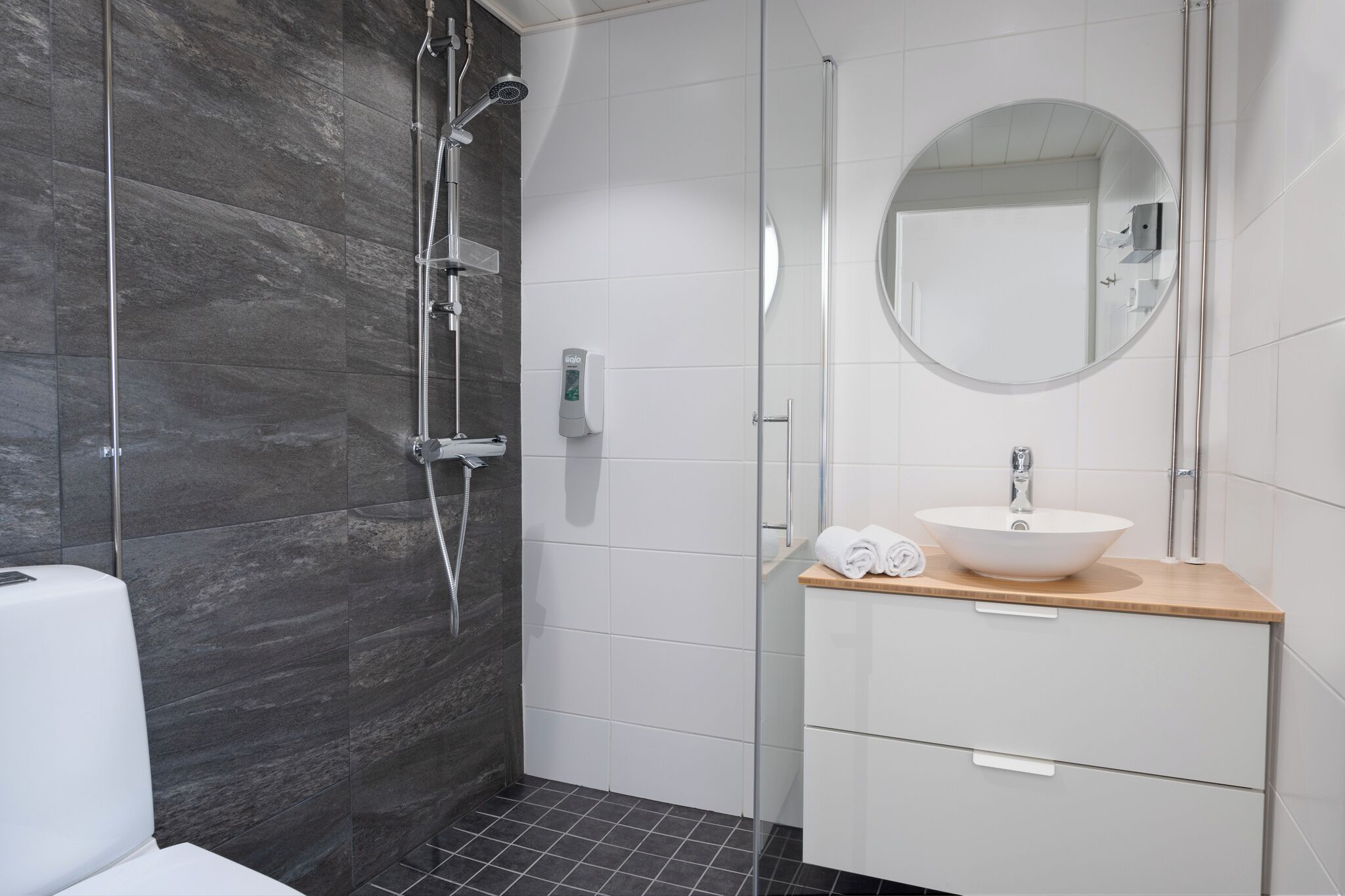 Un baño moderno con cabina de ducha de vidrio, un lavamanos blanco y encimera de madera, y un espejo redondo.