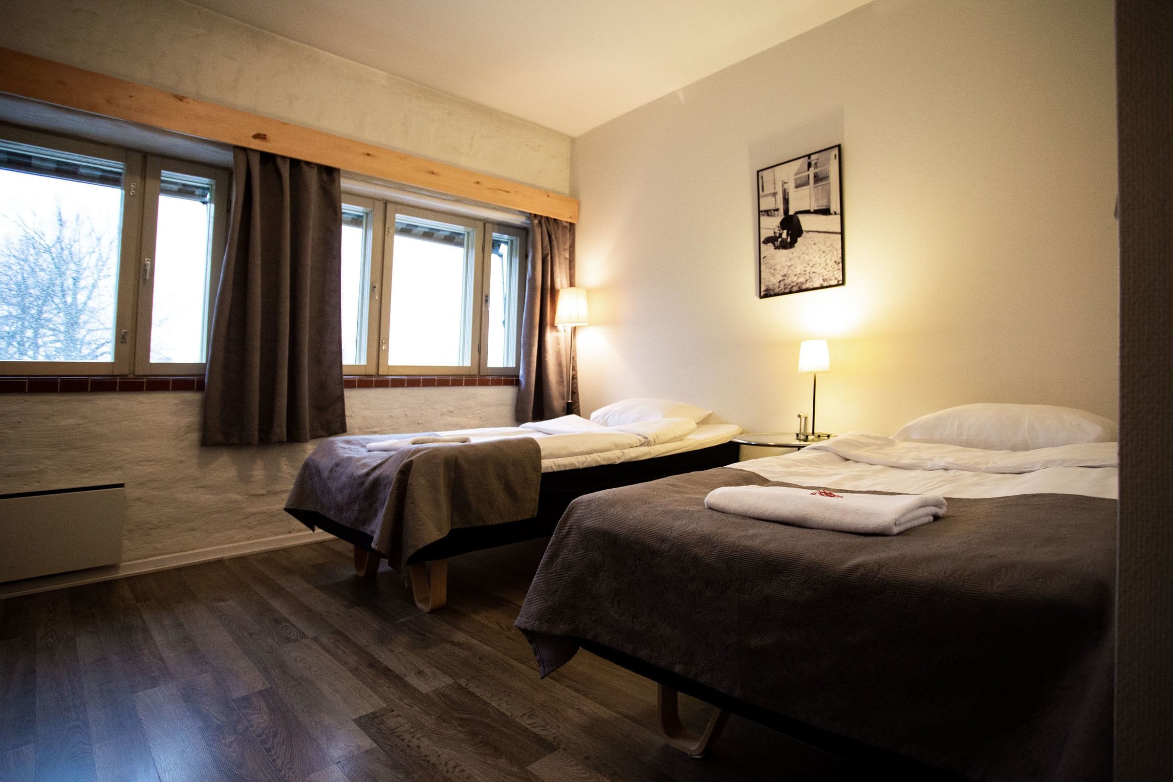 Habitació d'hotel amb dos llits, un sòl de fusta, una finestra amb cortines, una làmpada penjada a la paret i un quadre emmarcat.
