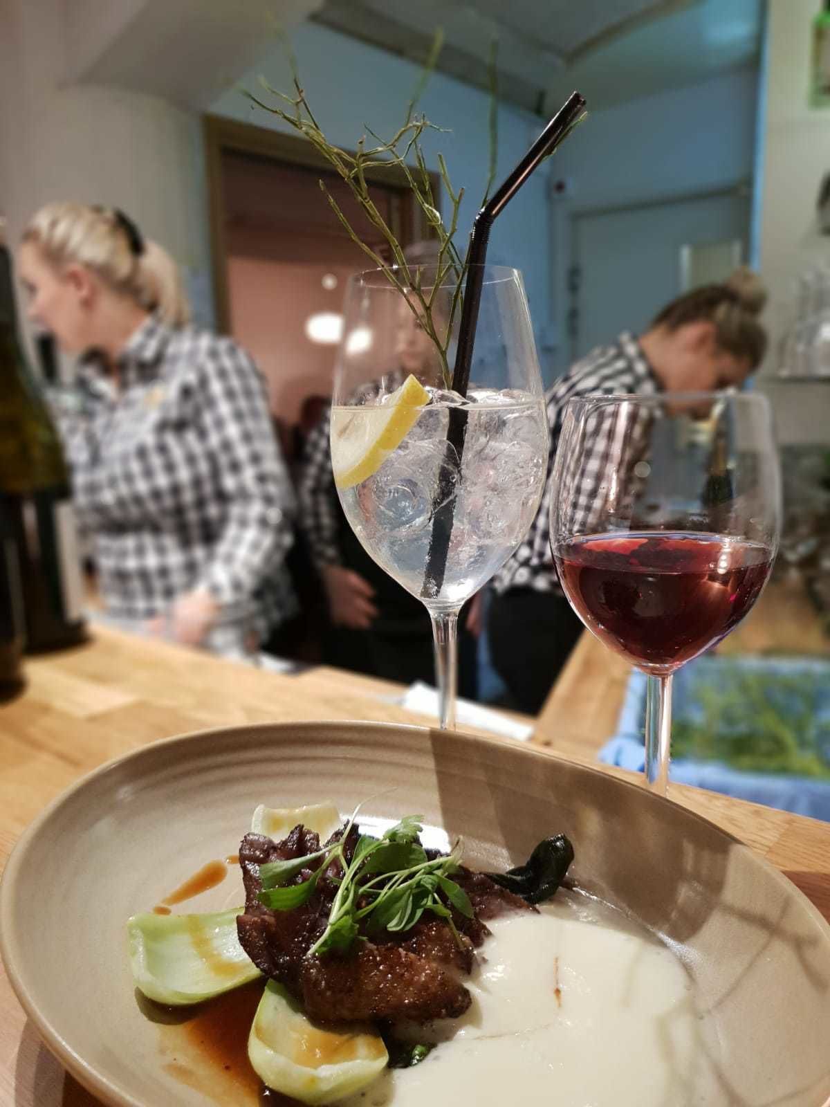 Una mesa de comedor con un plato de comida, dos vasos de bebidas y una zona de bar con gente al fondo.