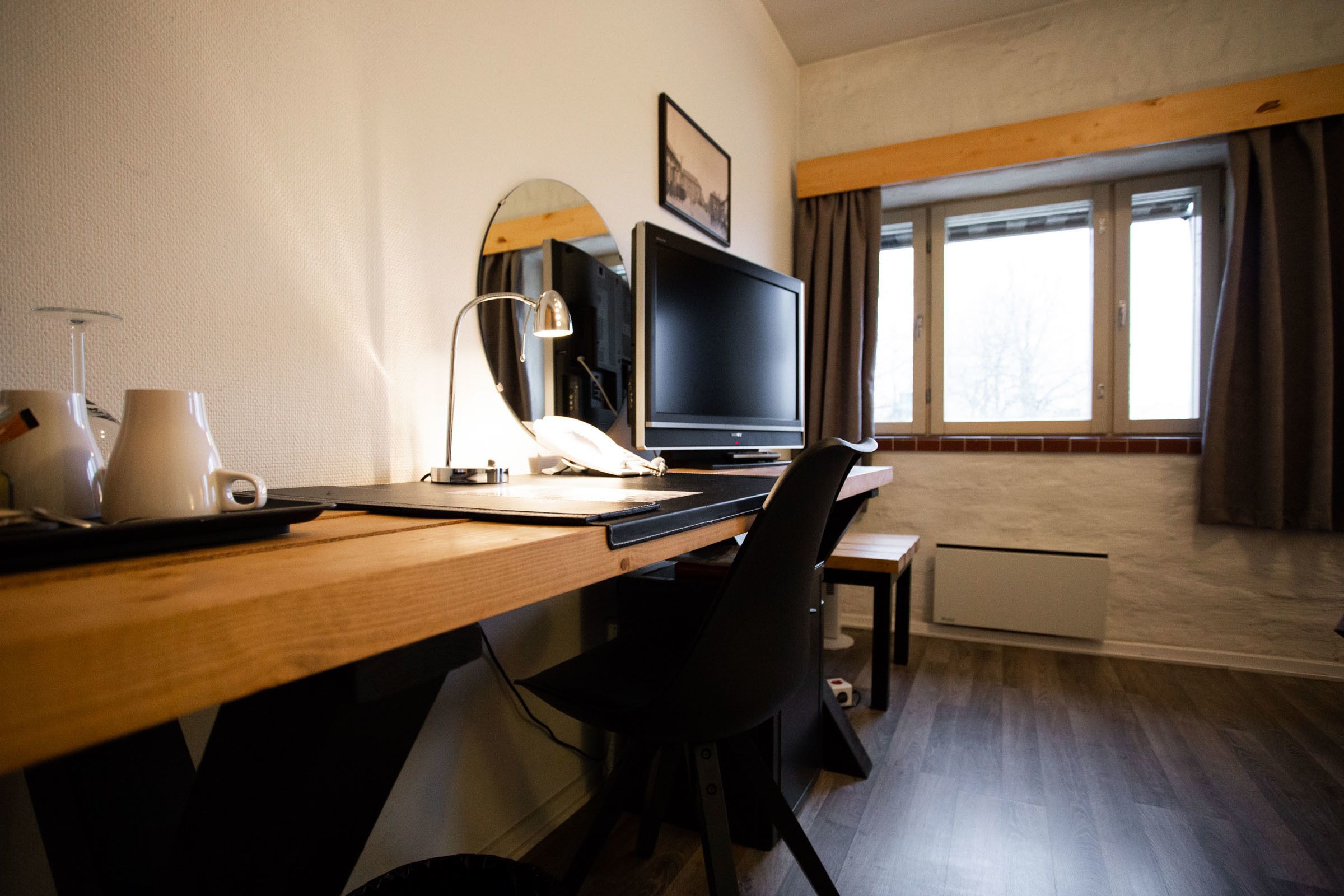 A hotel room with a wooden desk, a television, a lamp, a chair, and a window with curtains.