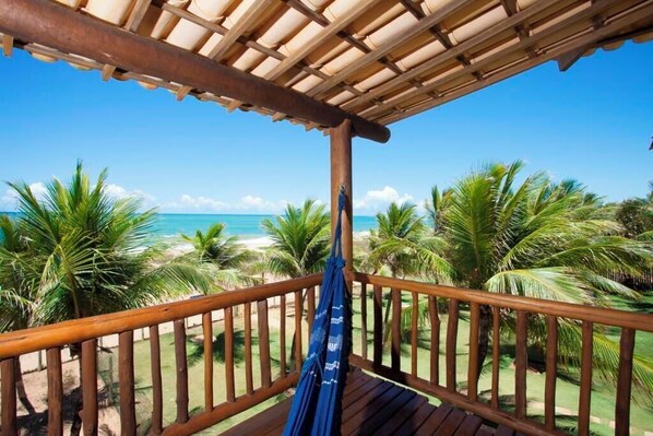 Una terraza de madera con una hamaca, palmeras y vista a la playa y el océano.