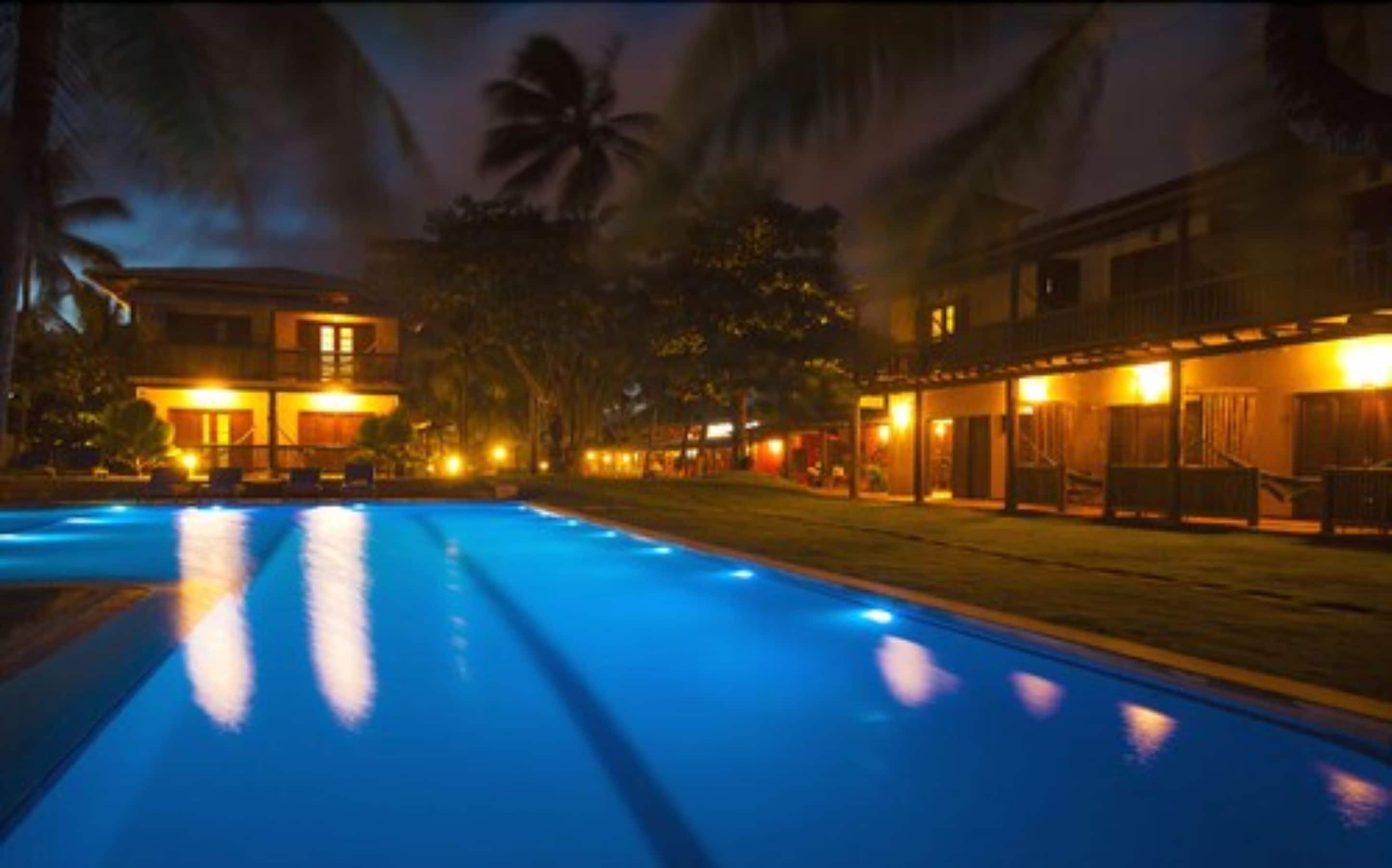 A nighttime view of a hotel with a swimming pool, palm trees, and illuminated buildings.