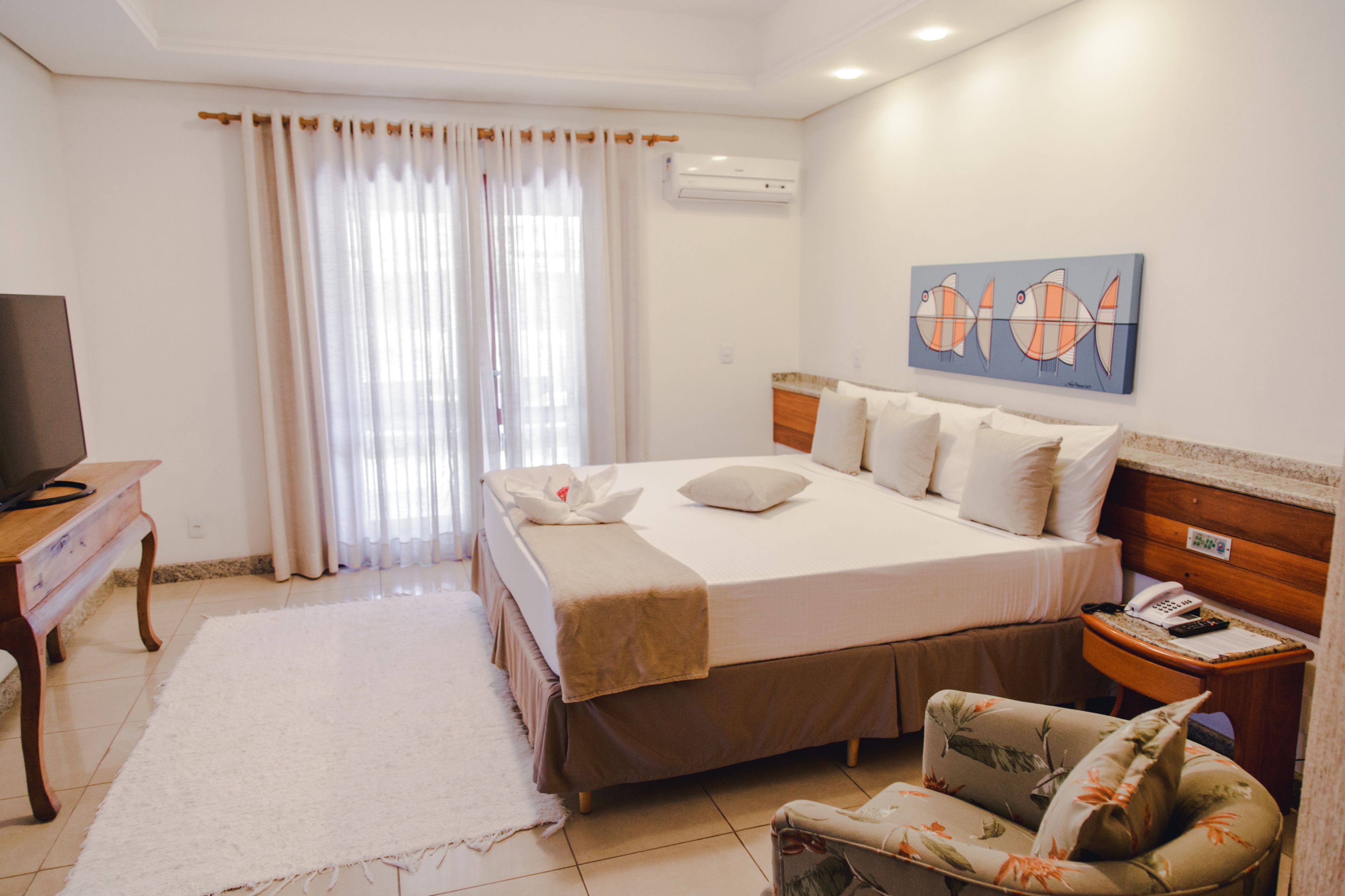 Una habitación de hotel con una cama, un televisor, una silla, un cuadro y una ventana con cortinas.