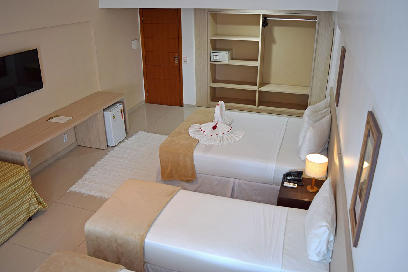 Habitación de hotel con dos camas, televisión, un refrigerador pequeño, mesita de noche con lámpara y armario.
