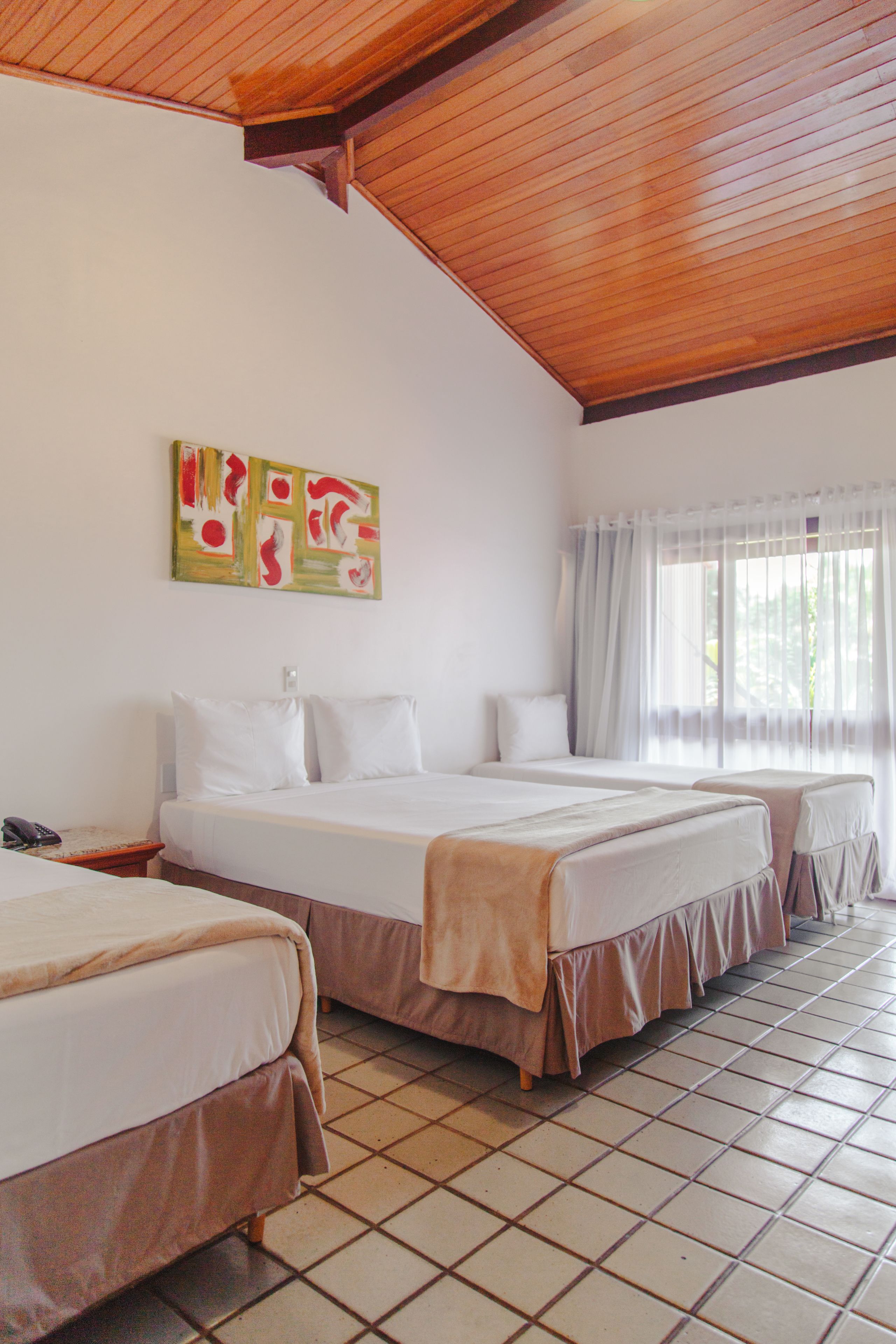 A hotel room with two beds, a wooden ceiling, a tiled floor, and a window with curtains.