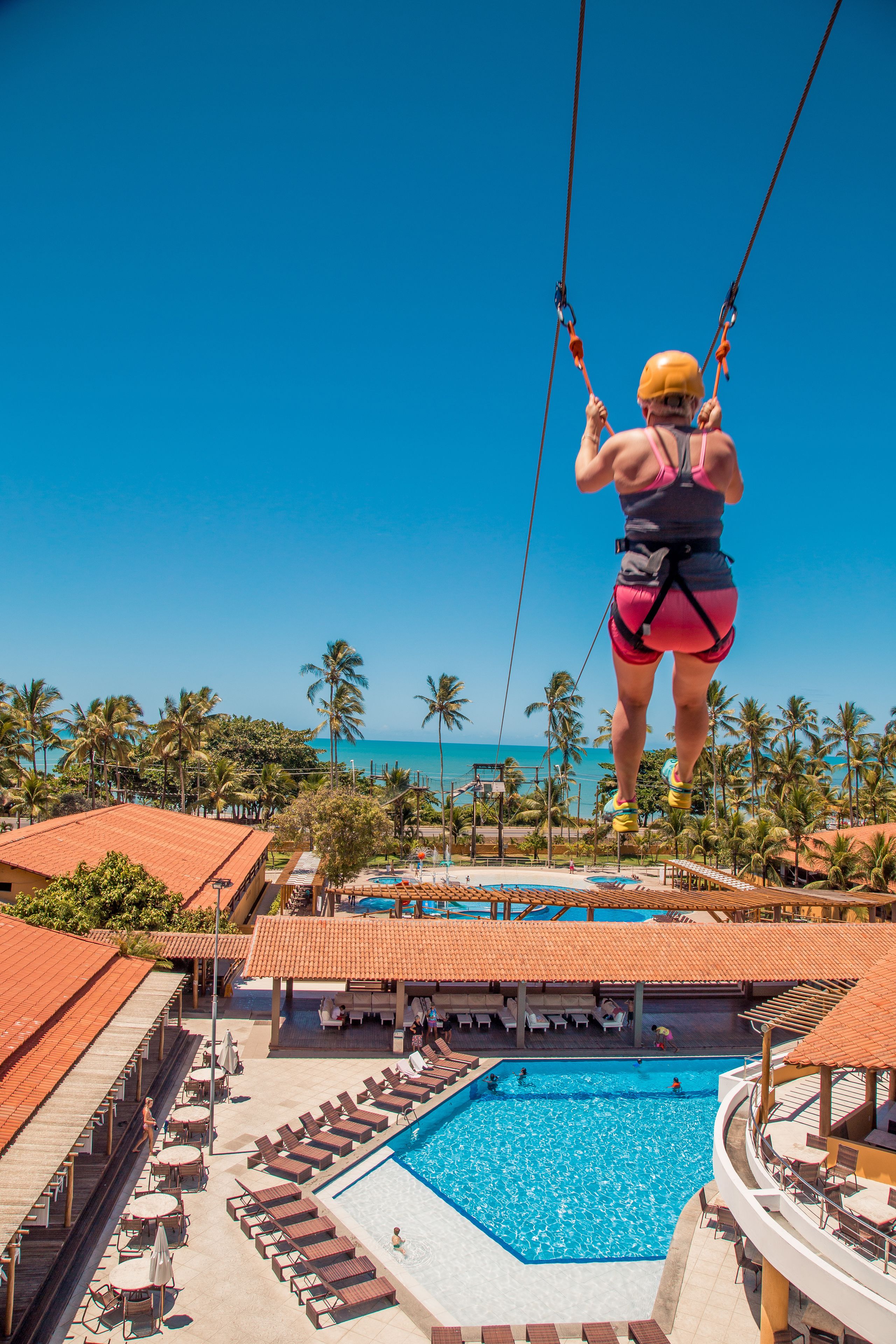 Uma pessoa faz bungee jumping de uma plataforma alta, com uma piscina e cadeiras de lounge abaixo.