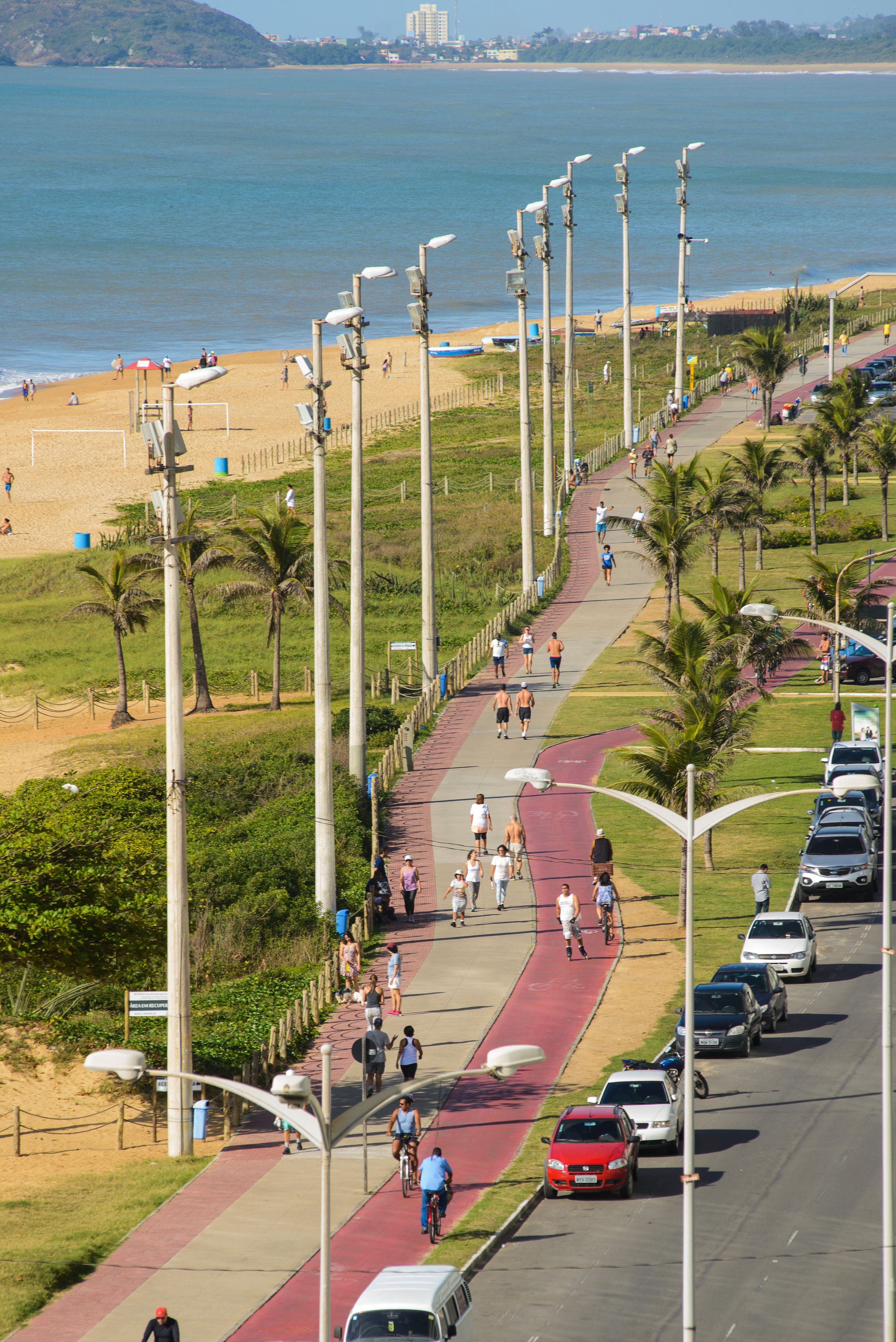 A coastal road with a red bike path, parked cars, and a beach in the background.