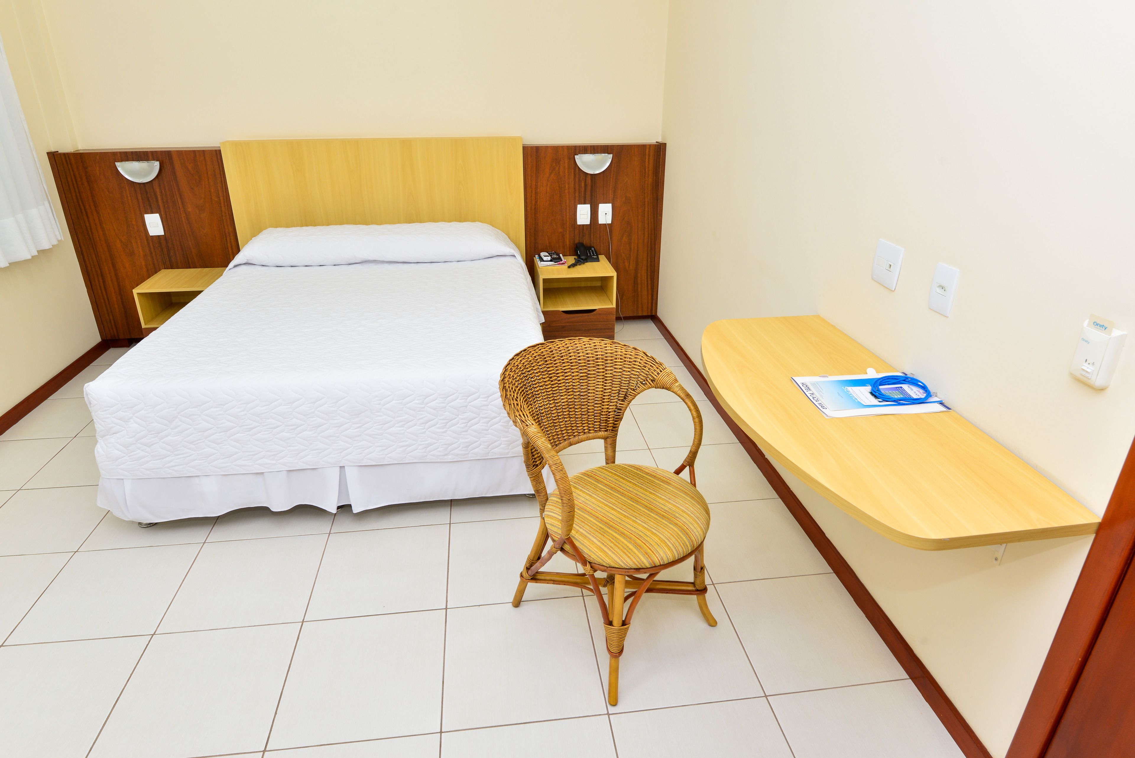 Habitación de hotel con cama, mesita de noche de madera, silla de mimbre, escritorio de madera y toma de corriente.