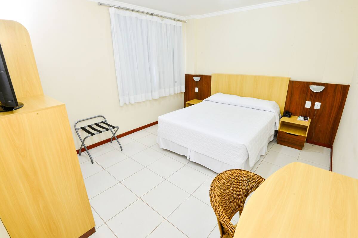 Ein Hotelzimmer mit einem Bett, einem kleinen Tisch, einem Stuhl, einem Fernseher und einem Fenster mit Vorhängen.