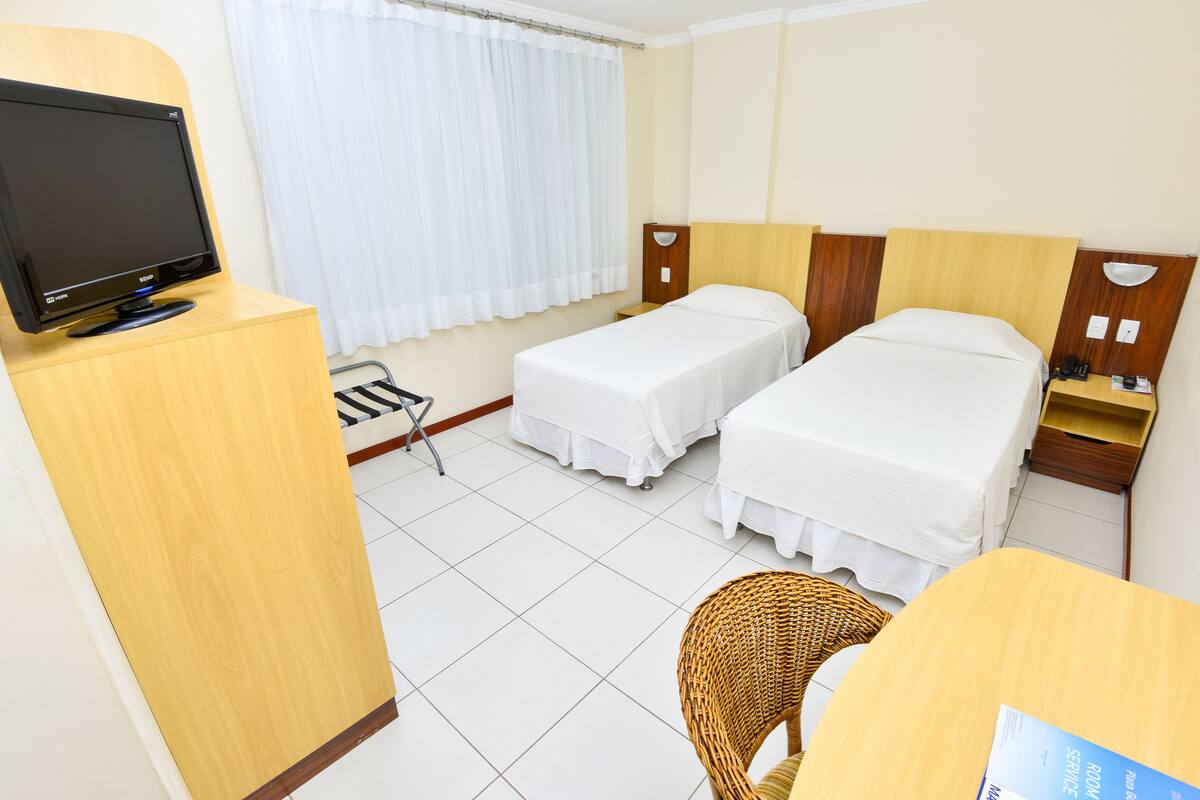 Ein Hotelzimmer mit zwei Einzelbetten, einem Fernseher auf einem Holzboden, einem Korbstuhl und einem Schreibtisch mit Stuhl.