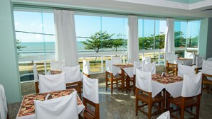 Un restaurante con mesas dispuestas para comer, grandes ventanales con vistas al océano y un balcón con vista a la playa.