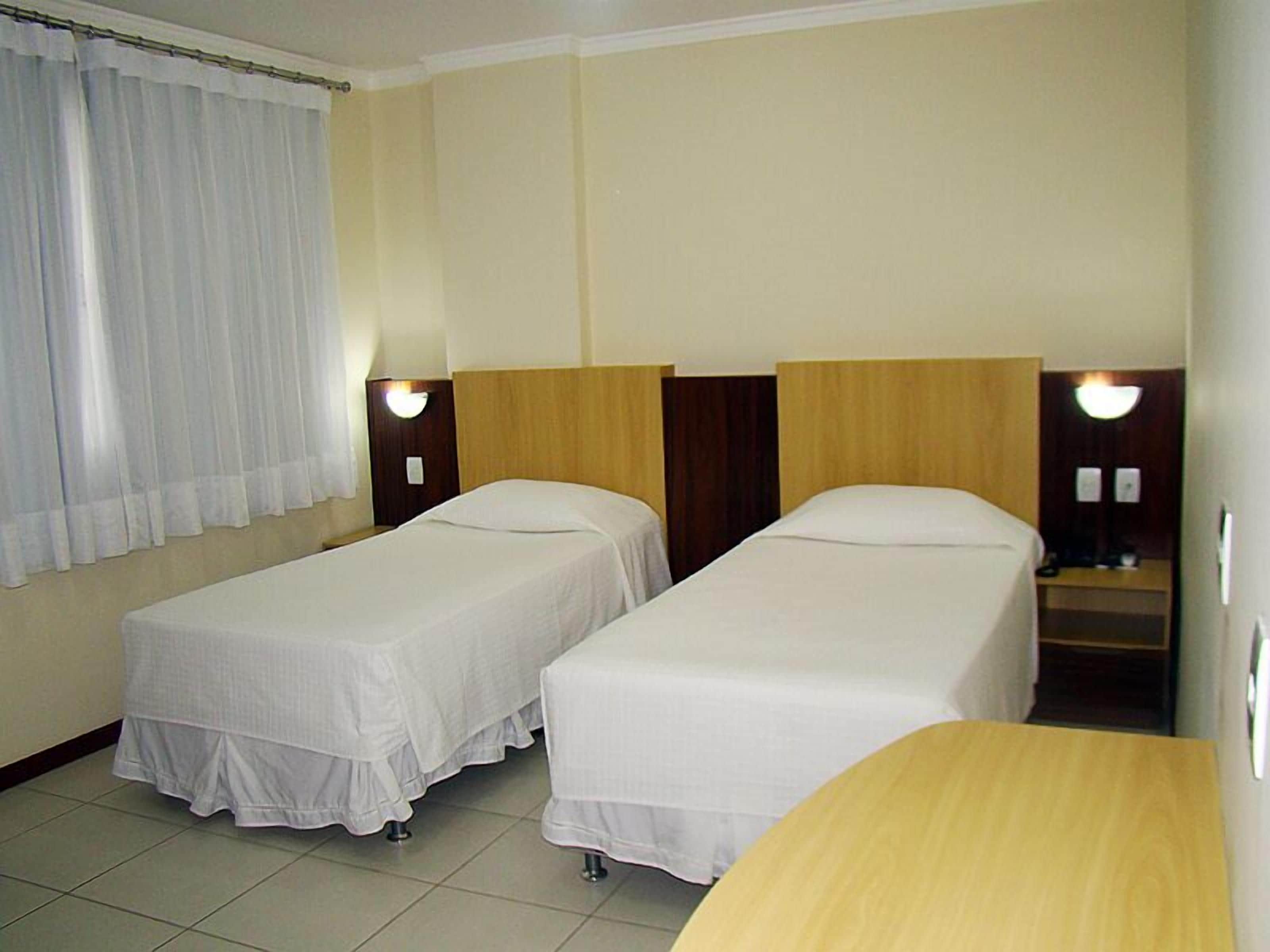 Quarto de hotel com duas camas de solteiro, uma mesa de cabeceira de madeira, uma mesinha e uma janela com cortinas.