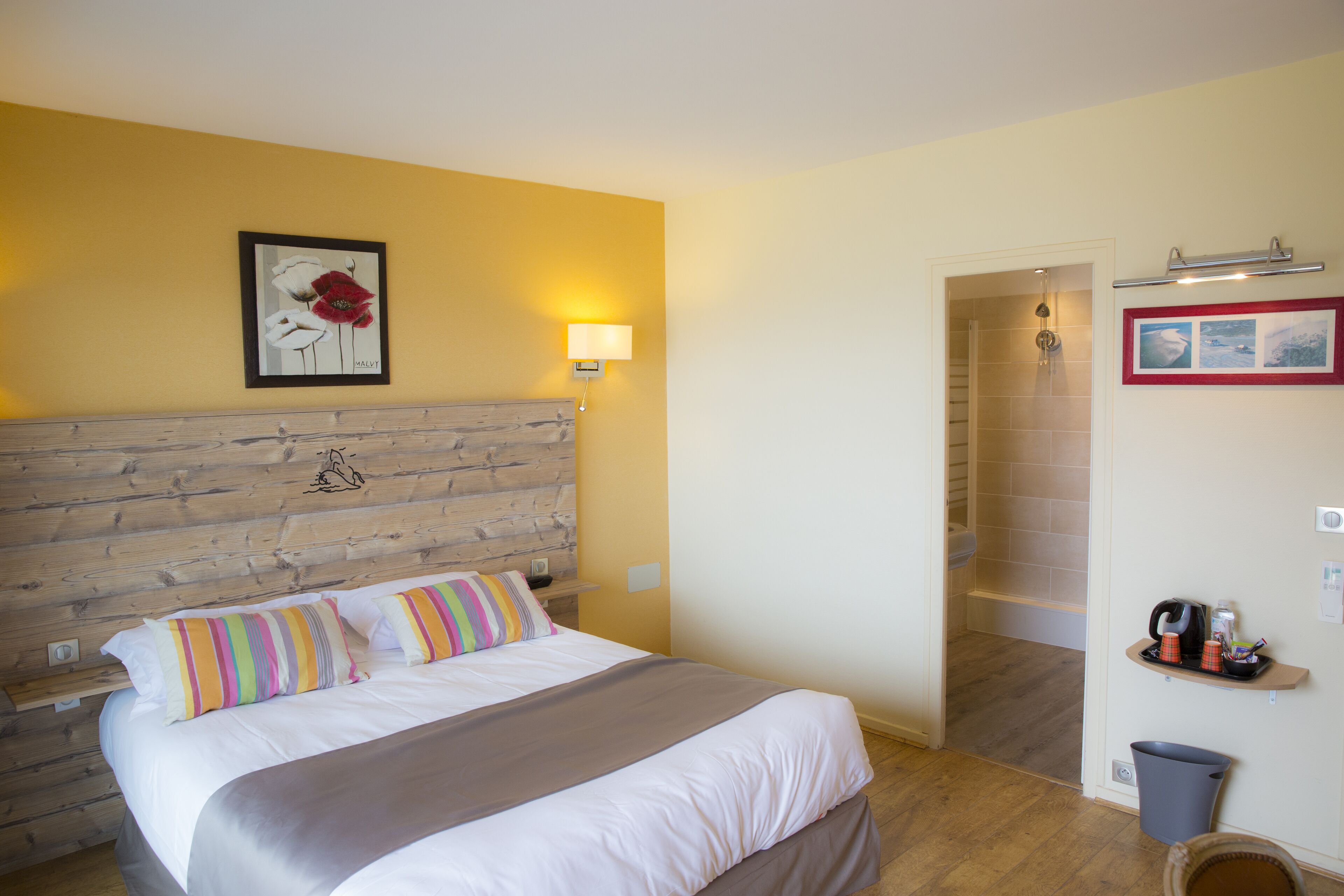 A bedroom with a wooden headboard, a bed with striped pillows, a nightstand, a wall-mounted lamp, a framed picture, a shower area, and a shelf with toiletries.