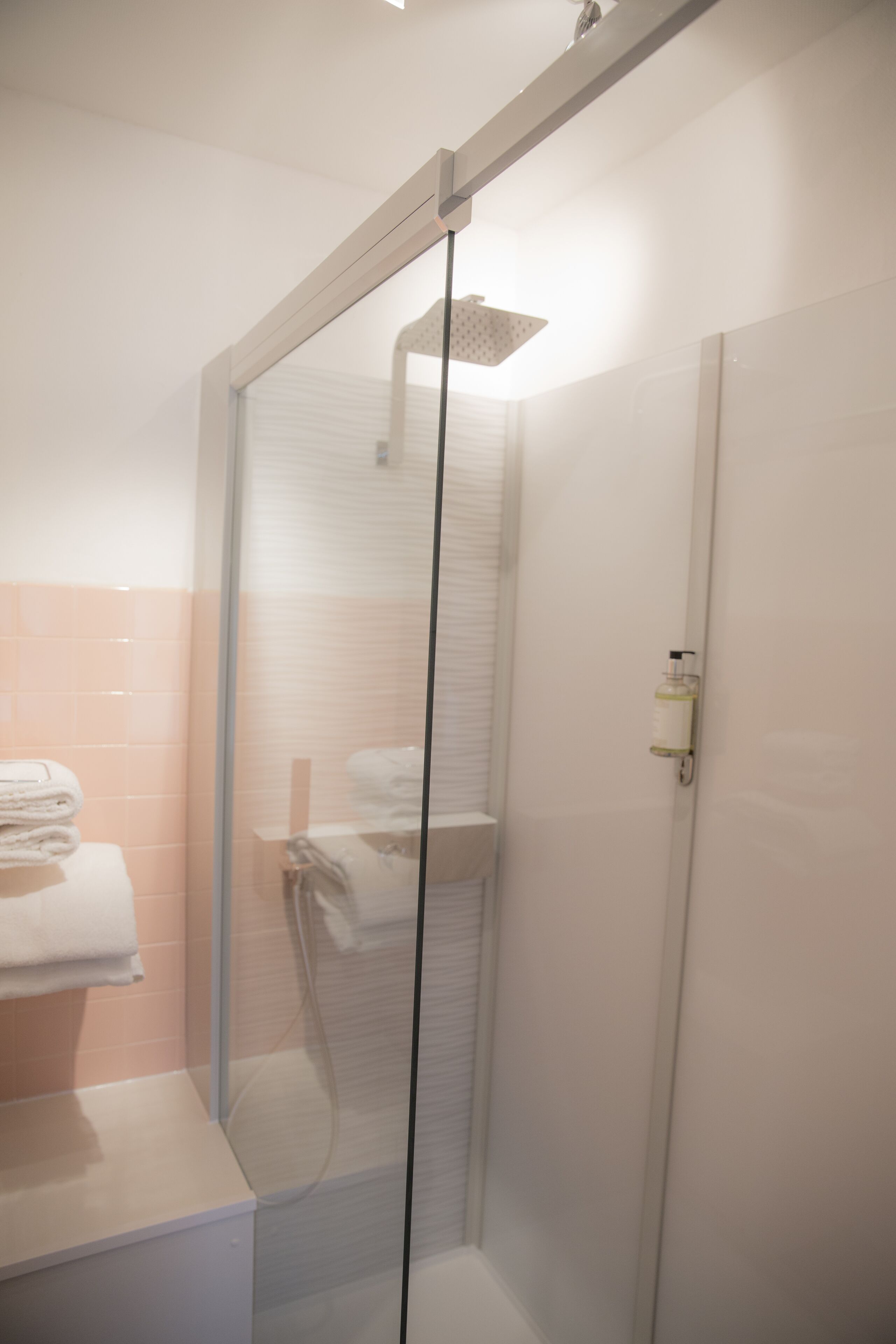 Une douche fermée par des parois de verre, avec un mur recouvert de carreaux blancs et des toilettes blanches visibles sur la gauche.