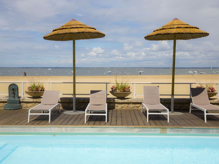 A poolside area with lounge chairs and thatched umbrellas, overlooking a beach.
