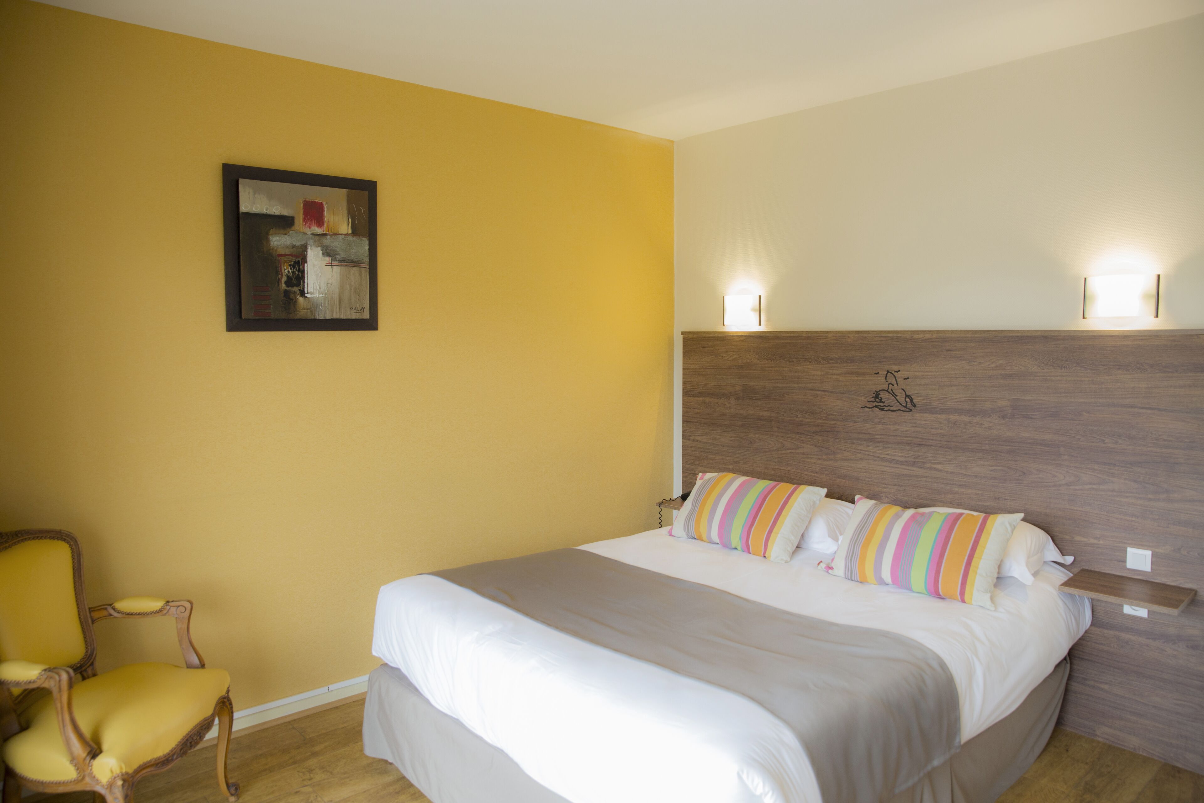 A hotel room with a bed, a chair, a painting on the wall, and two wall-mounted lights.
