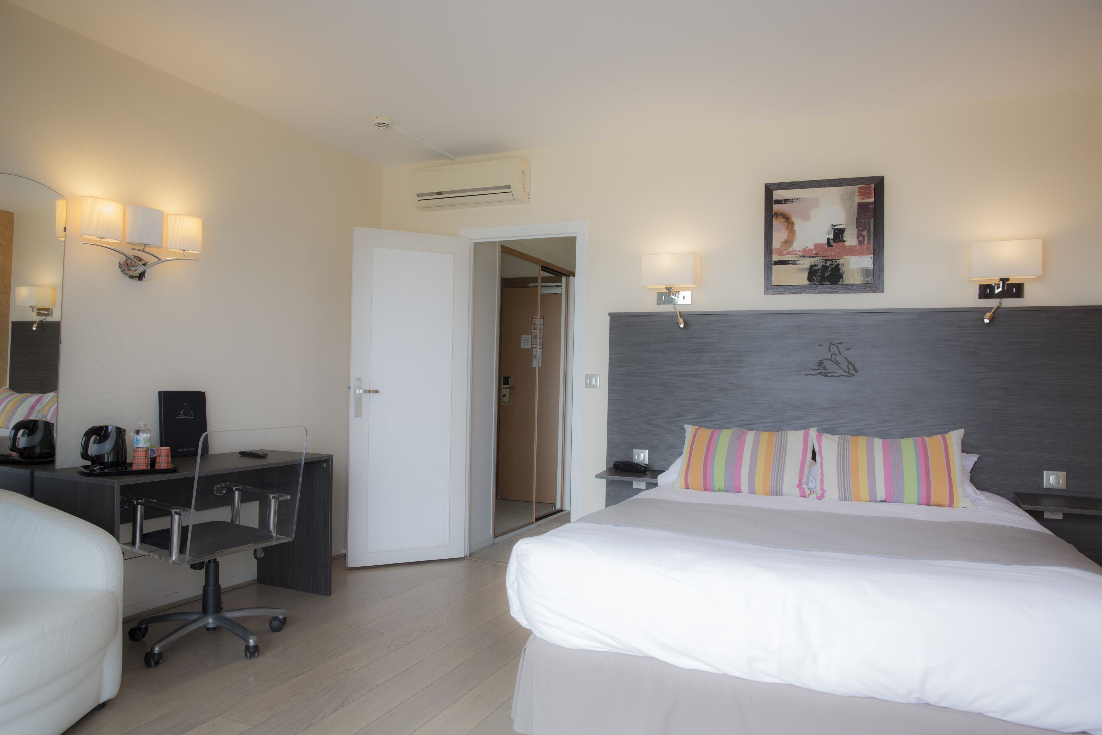 A hotel room with a bed, a desk, a chair, a nightstand, and a wall-mounted lamp.