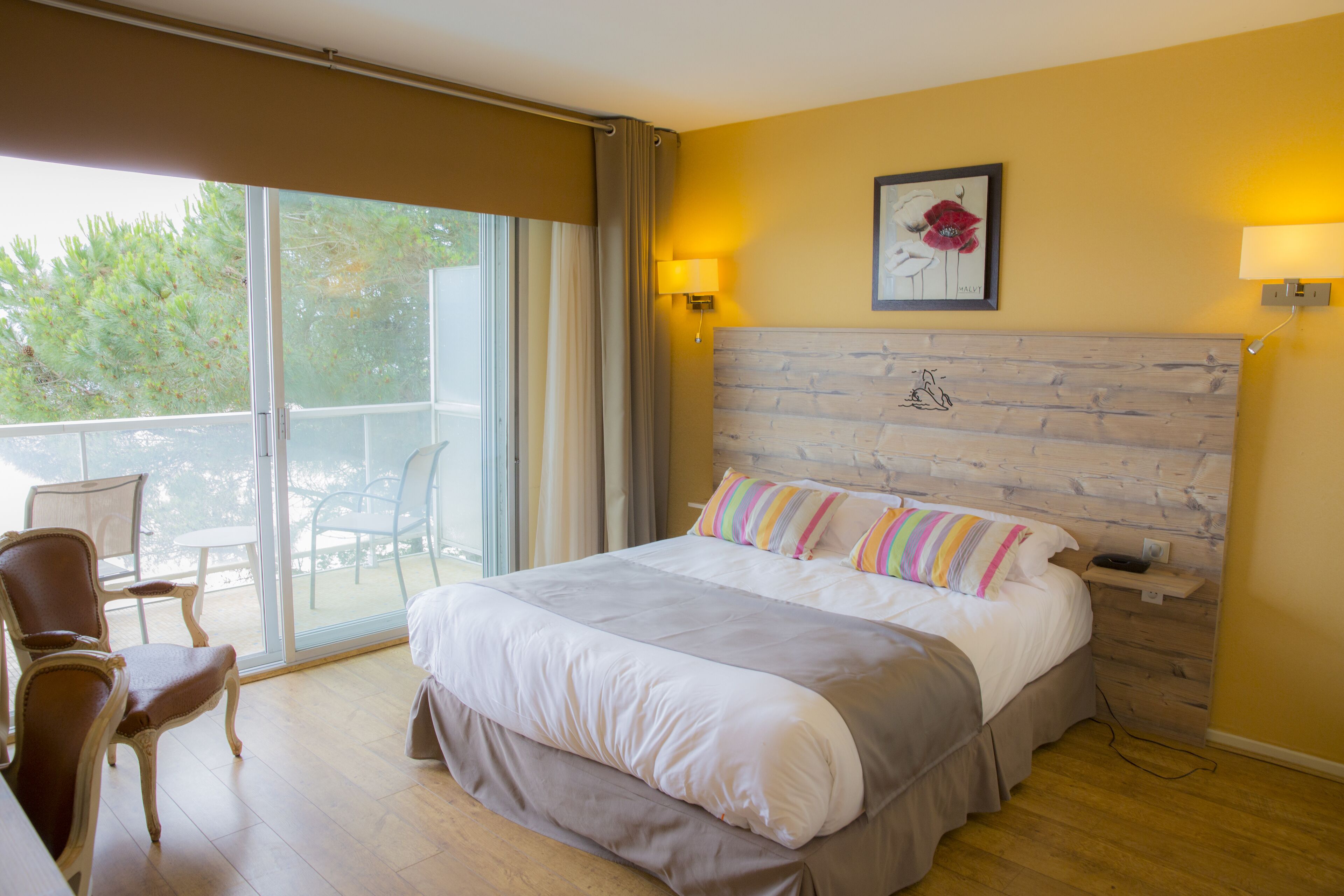 A hotel room with a large bed, a wooden headboard, a balcony with chairs, and a painting on the wall.