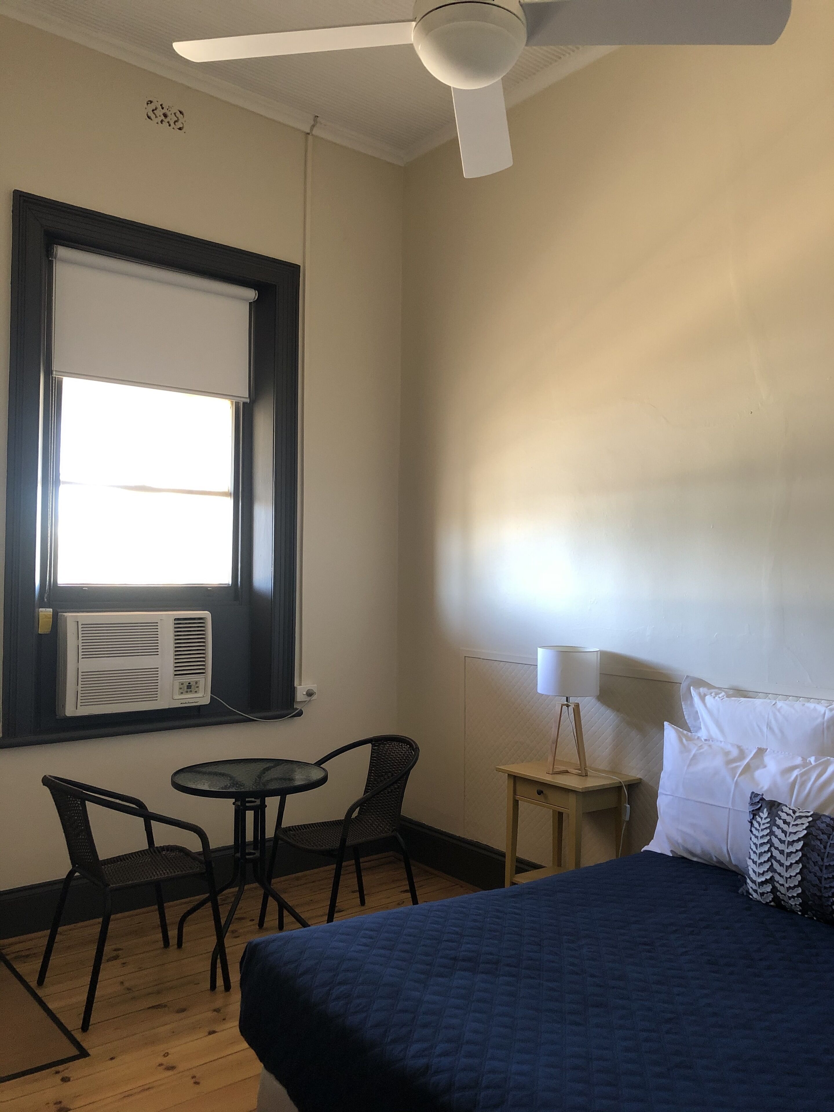 A bedroom with a bed, a ceiling fan, a window with blinds, a table with chairs, and a bedside lamp.