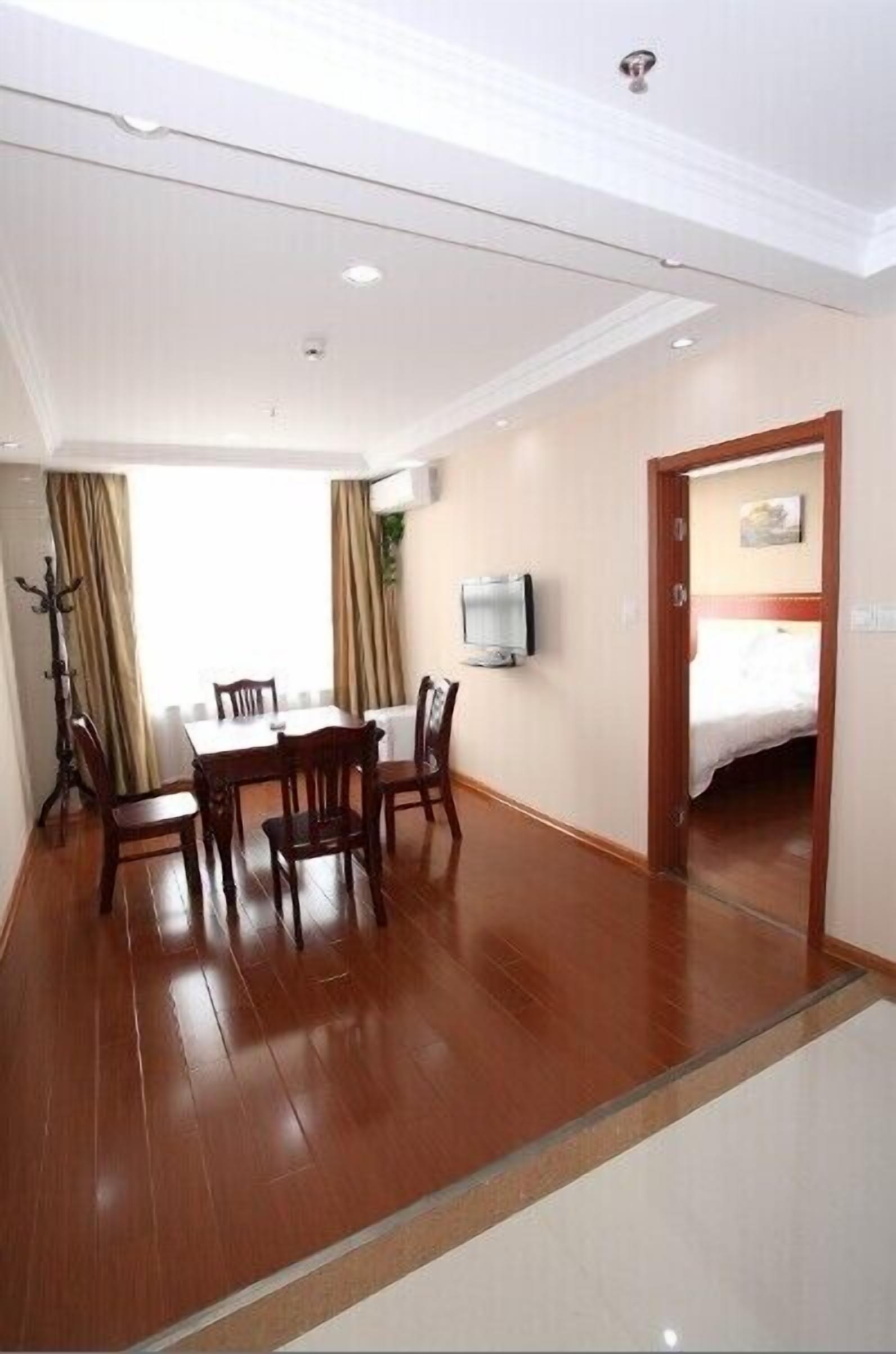 A hotel room with a wooden floor, a dining table with chairs, a flat-screen TV mounted on the wall, and a bed in the adjacent room.