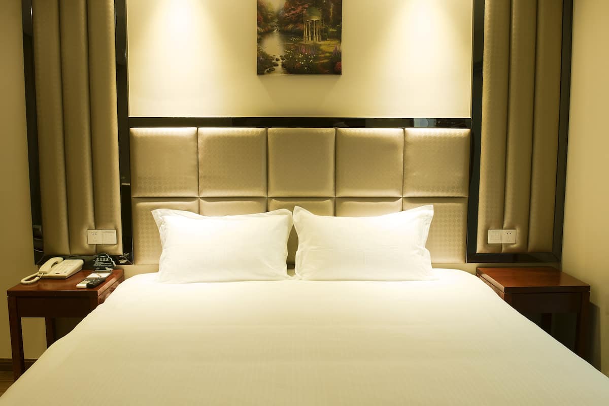 A hotel room with a large bed, two pillows, a headboard, a bedside table with a phone and remote control, and a painting on the wall.