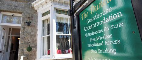Un cartel verde para 'The Dunedin Guesthouse' que enumera comodidades como todas las habitaciones, baño privado, wifi gratuito, acceso a banda ancha y no fumadores.