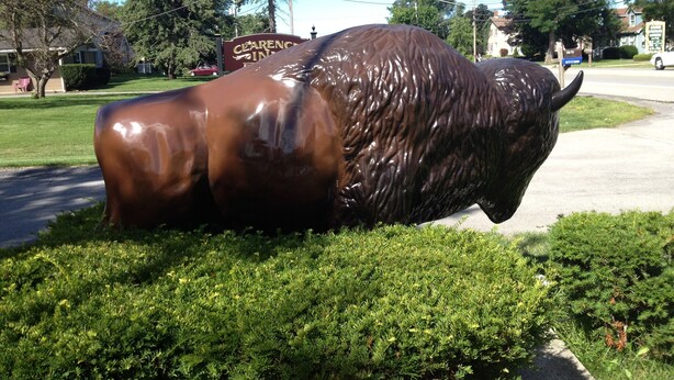 A bronze bull sculpture in a grassy area with a road and a sign for 'CLEANERS INN' in the background. - Clarence Inn (Clarence)