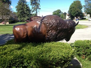 Una escultura de bronce de un toro en un área con césped, con una carretera y un letrero que dice 'CLEANERS INN' al fondo.