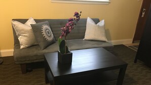 A living room with a dark wooden coffee table, a grey sofa with patterned cushions, and a potted plant with purple flowers. - Clarence Inn (Clarence)