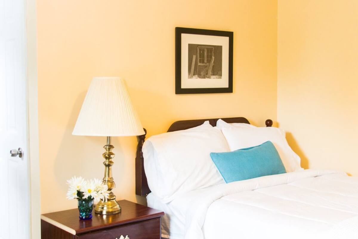 A neatly made bed with white linens and a blue pillow, a bedside lamp, and a small vase with flowers on a nightstand.