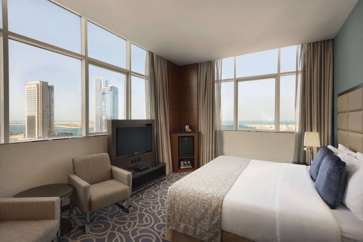 A hotel room with a large bed, a TV, a seating area, and a view of tall buildings.