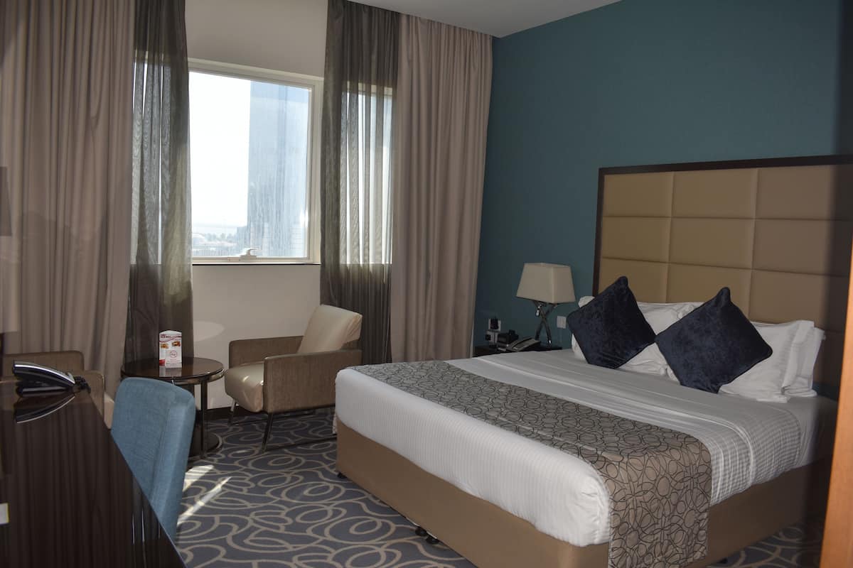 A hotel room with a bed, a desk, a chair, a nightstand, and a window with curtains.