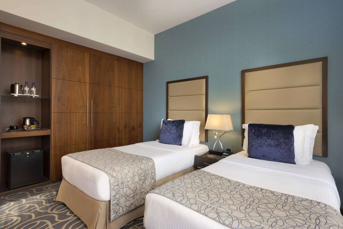 A hotel room with two beds, a wardrobe, a desk with a kettle, and a mini fridge.